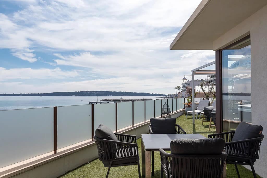 Where are the Best Neighbourhoods to Live in Cannes? 6 Image of a modern terrace in Palm Beach Cannes with outdoor seating and glass railings, overlooking a calm sea under a partly cloudy sky.