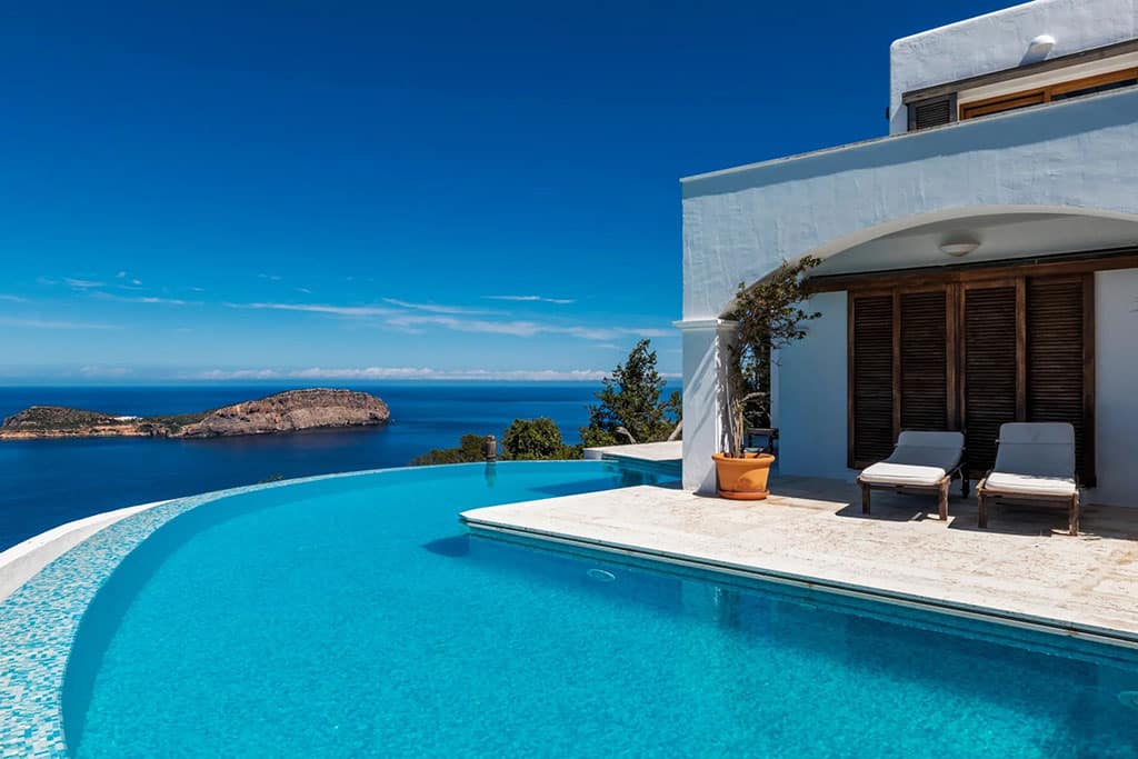 Find Your Dream Villa in Ibiza’s Most Prestigious Locations 3 Image of a luxurious seaside villa in Ibiza with an infinity pool overlooking a calm blue ocean and a distant rocky island under clear skies.