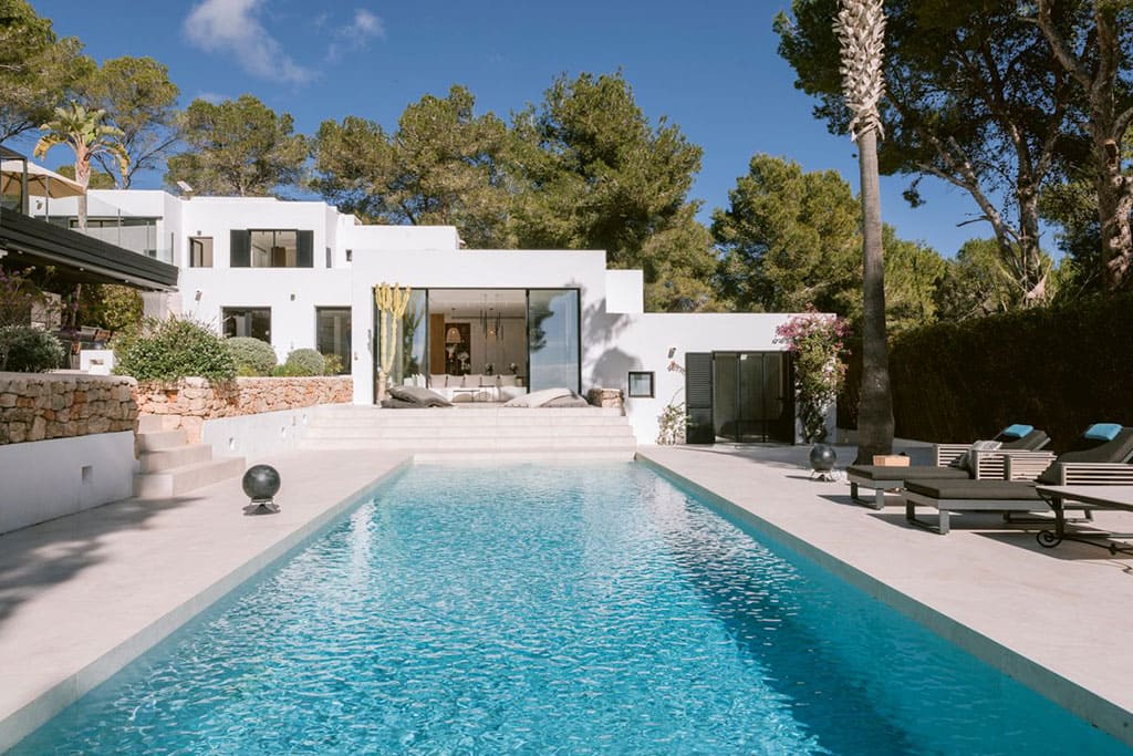Find Your Dream Villa in Ibiza’s Most Prestigious Locations 4 Image of a modern white villa in Ibiza with a long rectangular pool, surrounded by trees, sun loungers, and a peaceful garden setting.