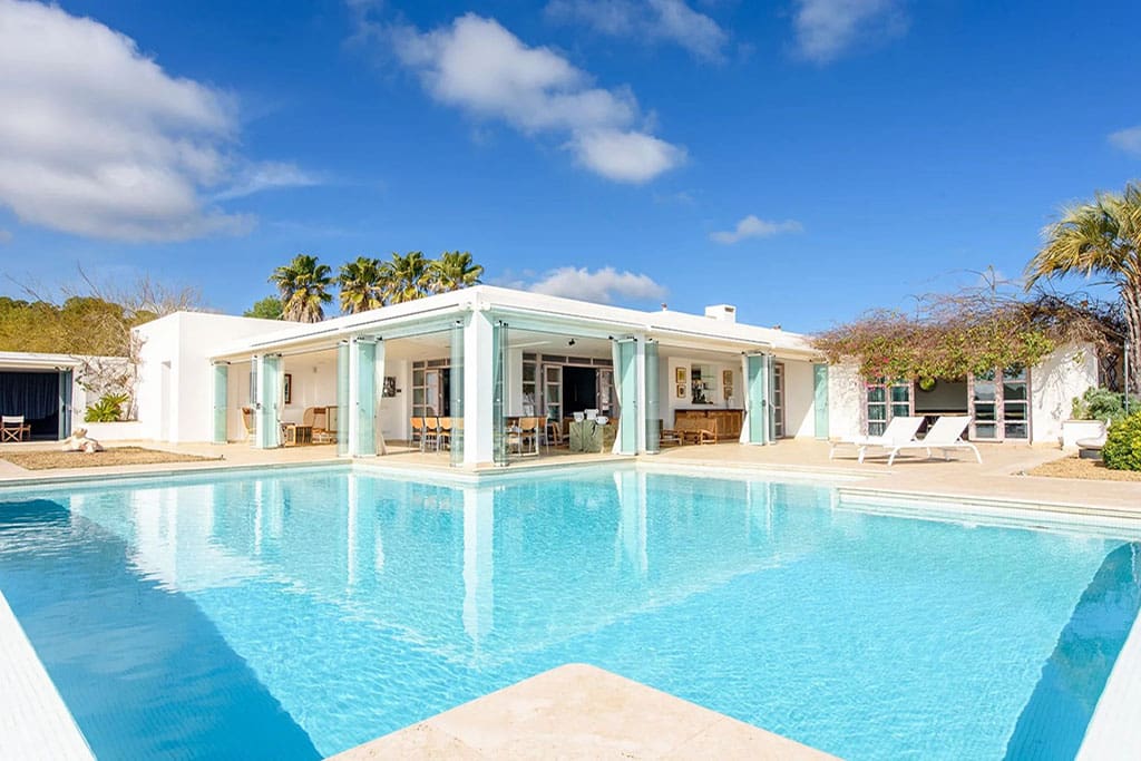 Find Your Dream Villa in Ibiza’s Most Prestigious Locations 7 Image of a bright modern villa in Roca Llisa, Ibiza with glass doors opening onto a large turquoise pool, surrounded by palm trees and clear blue skies.