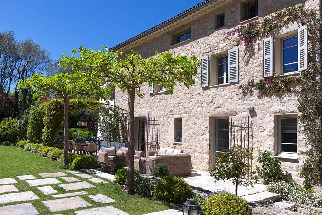 Buyers Guide to the Most Luxurious Properties in Valbonne 6 Image of a rustic stone villa with shuttered windows and a shaded patio featuring outdoor seating surrounded by lush greenery and manicured gardens.