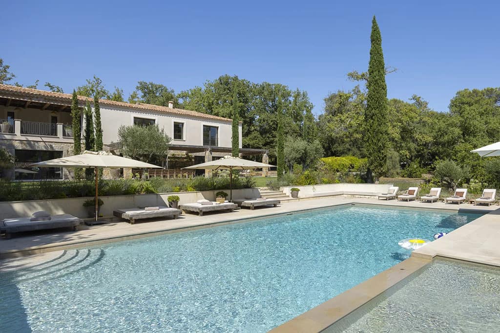 Buyers Guide to the Most Luxurious Properties in Valbonne 3 Image of a large outdoor swimming pool surrounded by lounge chairs and umbrellas beside a Mediterranean-style villa under a clear blue sky.