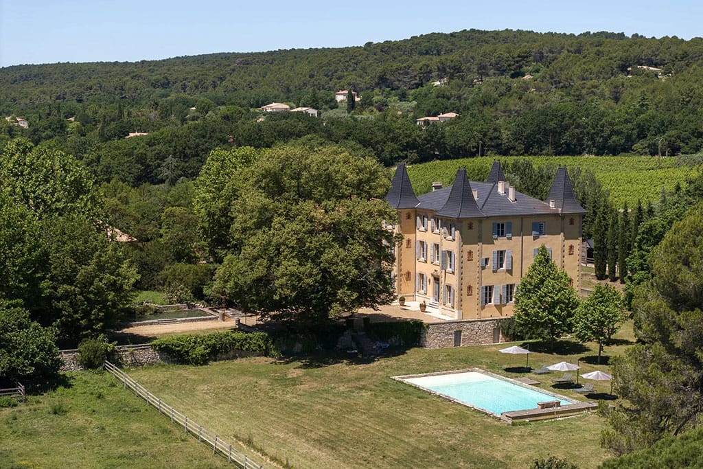 Image of a grand stone château in Provence surrounded by lush greenery, with towers, arched windows, and a clear blue sky overhead.