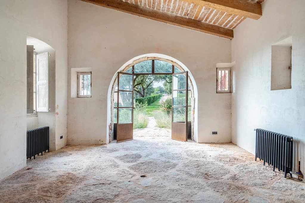Image of a rustic interior with stone flooring, wooden ceiling beams, and arched glass doors opening to a garden with trees and greenery.