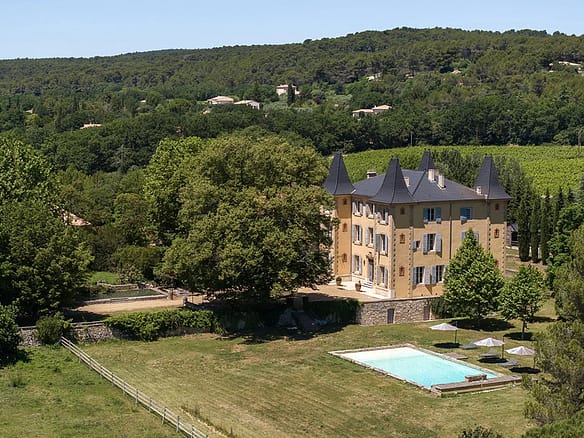 Image of a grand stone château in Provence surrounded by lush greenery, with towers, arched windows, and a clear blue sky overhead.