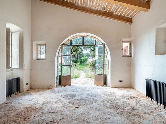 Image of a rustic interior with stone flooring, wooden ceiling beams, and arched glass doors opening to a garden with trees and greenery.