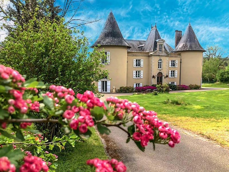 Image of a grand luxury French château with tall turrets and elegant stone architecture, surrounded by manicured lawns, dense trees, and a bright partly cloudy sky.