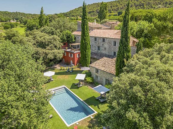 Image of a secluded stone chateau surrounded by lush trees, featuring a rectangular outdoor pool, shaded lounge areas, and rolling green hills.