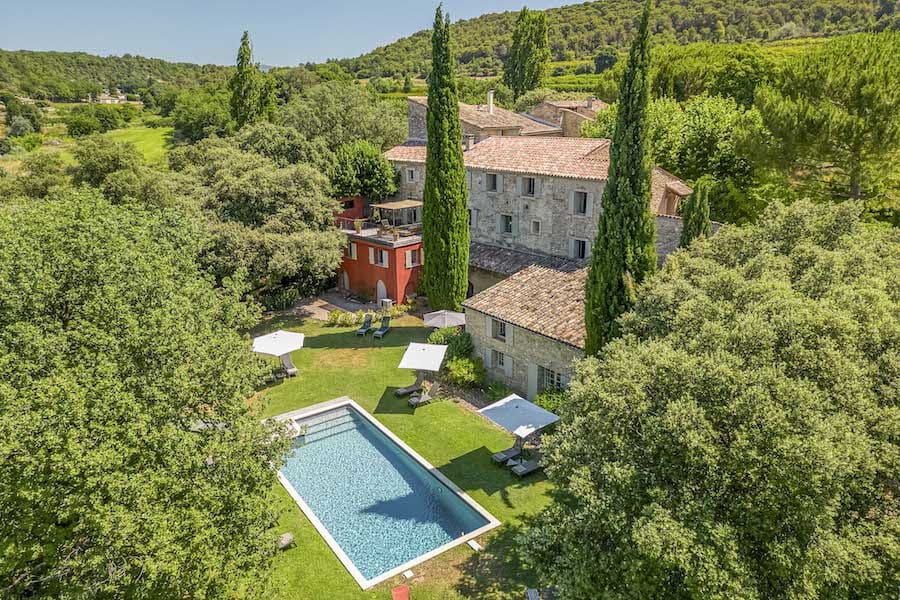 Image of a secluded stone chateau surrounded by lush trees, featuring a rectangular outdoor pool, shaded lounge areas, and rolling green hills.