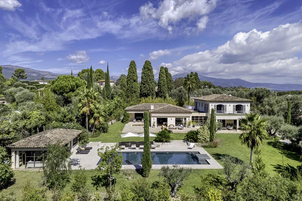 Buyers Guide to the Most Luxurious Properties in Valbonne 5 Image of a large villa surrounded by lush greenery and tall trees, featuring a central swimming pool and patio under a partly cloudy sky.
