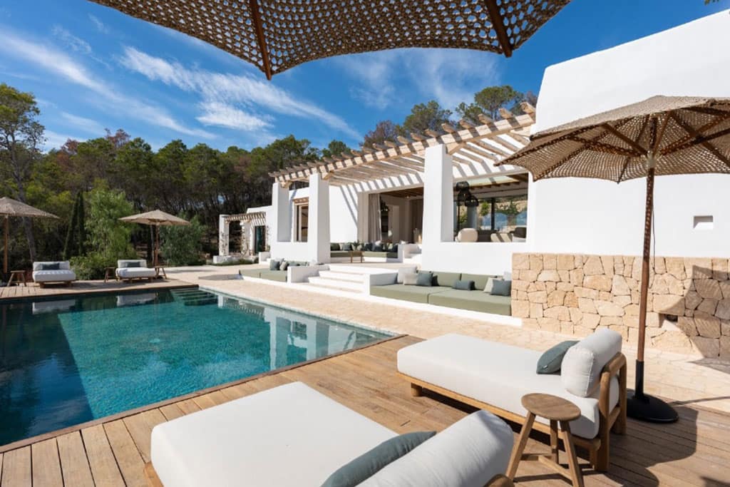 Find Your Dream Villa in Ibiza’s Most Prestigious Locations 5 Image of a stylish Mediterranean villa with white walls, stone accents, a wooden deck, and a serene pool surrounded by cushioned lounge seating.