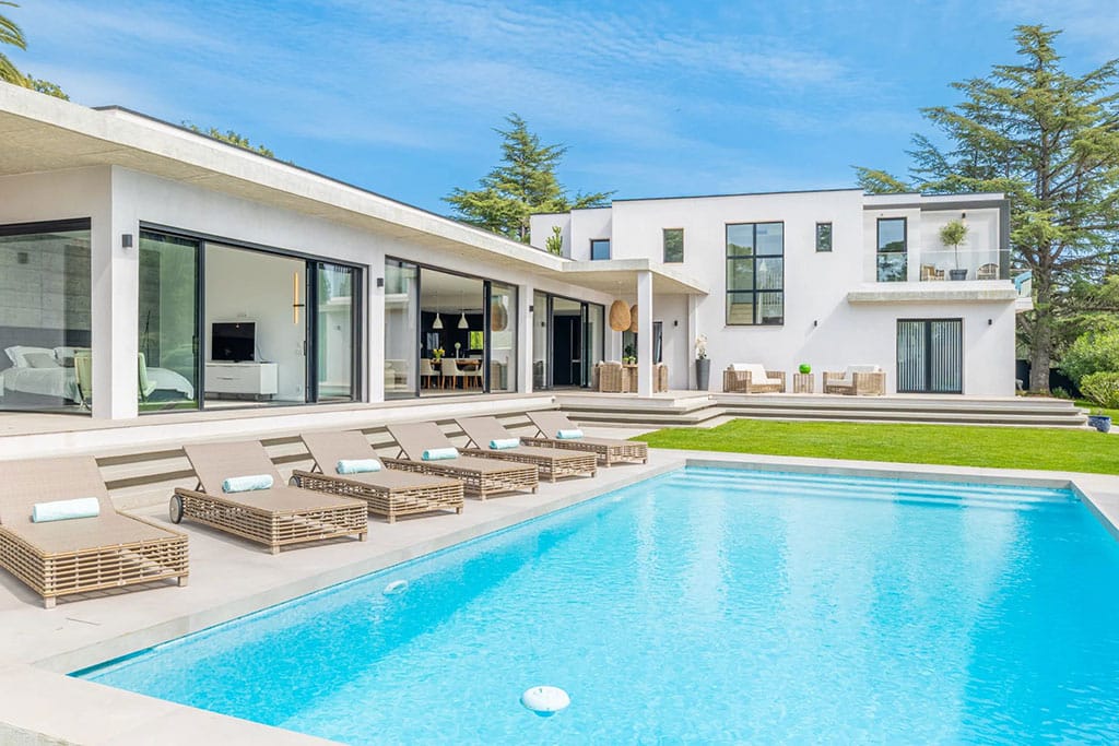 Buyers Guide to the Most Luxurious Properties in Valbonne 7 Image of a modern white villa with large glass doors opening to a poolside terrace lined with wicker lounge chairs under a bright blue sky.