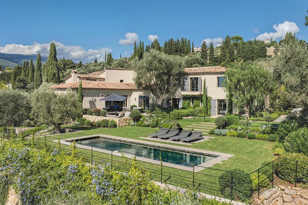Buyers Guide to the Most Luxurious Properties in Valbonne 4 Image of a Mediterranean-style villa surrounded by lush gardens and tall trees, featuring a rectangular swimming pool with lounge chairs on a manicured lawn.