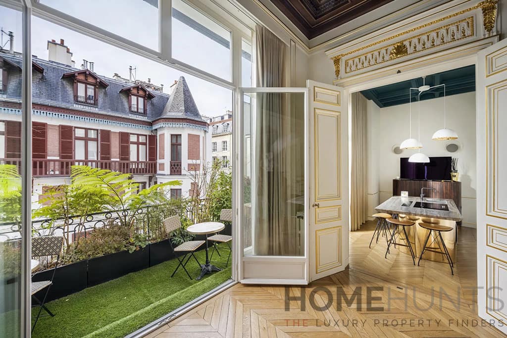 Looking to Buy in Paris? Discover 5 Incredible Luxury Apartments for Sale 2 Image of a stylish apartment with a small balcony featuring outdoor seating, gold-trimmed doors, and an elegant kitchen with pendant lighting.