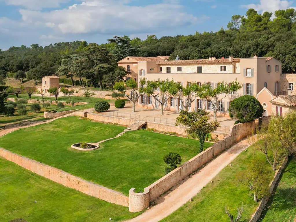 Is Provence Still a Safe Investment for International Buyers? 3 Image of a large estate in Provence, France, with manicured lawns, stone walls, tall windows, and tree-lined terraces set against a wooded hillside.